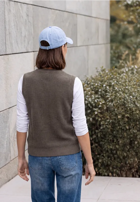 Person von hinten, weiße Kappe, weißes Langarmshirt, dunkle Weste, Jeans, vor Mauer und Sträuchern.