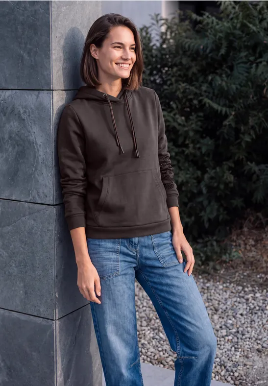Junge Frau in schwarzer Kapuzenjacke und Jeans lehnt lächelnd an einer grauen Wand im Freien.