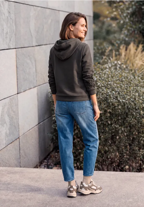 Frau in dunkler Kapuzenjacke und Jeans steht im Freien vor einer modernen Mauer.