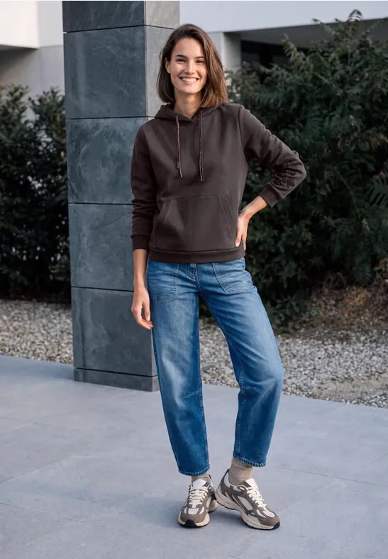 Junge Frau in Hoodie, Jeans und Sneakern vor modernem Gebäude, lächelnd.