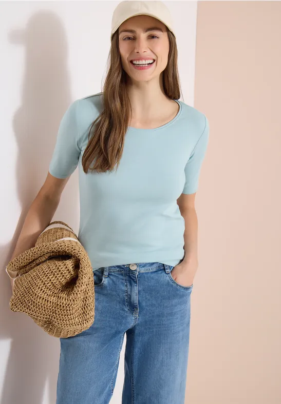 Frau in hellblauem Kurzarmshirt und Jeans hält eine Basttasche vor hellem Hintergrund.