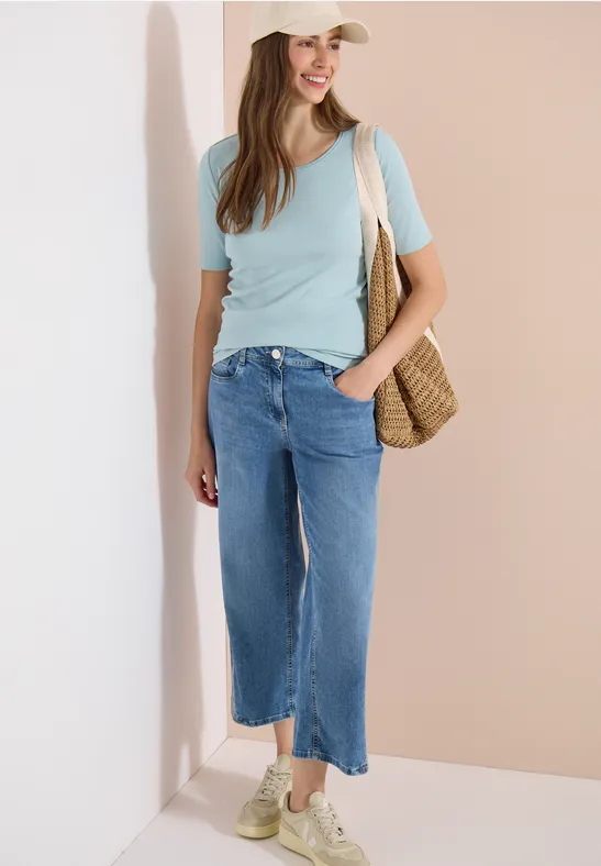 Frau in lockerer Sommerkleidung: hellblaues T-Shirt, weit geschnittene Jeans und Strohtasche lehnt an der Wand.