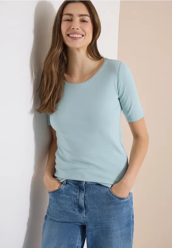 Frau lehnt an einer Wand, trägt ein hellblaues T-Shirt und Jeans.