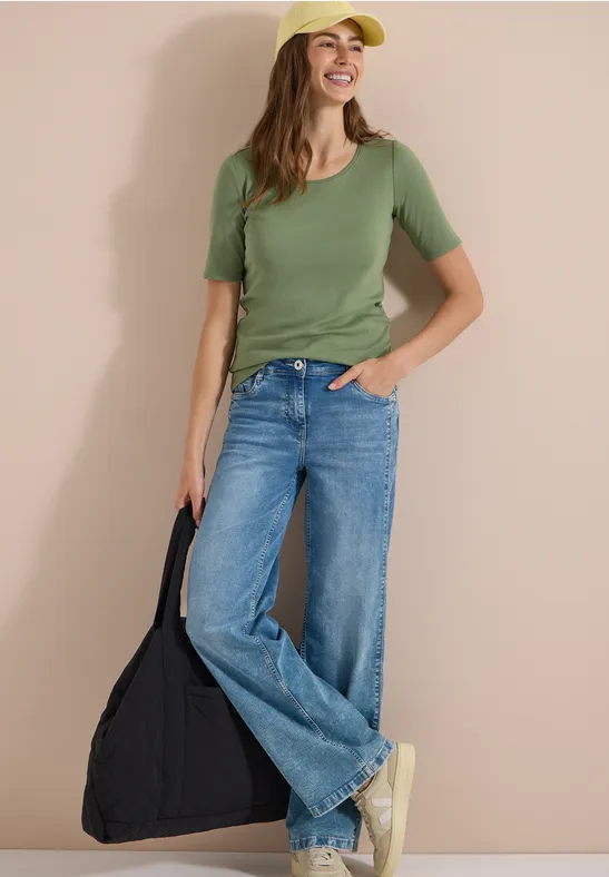 Vrouw in groen T-shirt, wijde jeans en witte sneakers met een zwarte tas tegen een neutrale achtergrond.