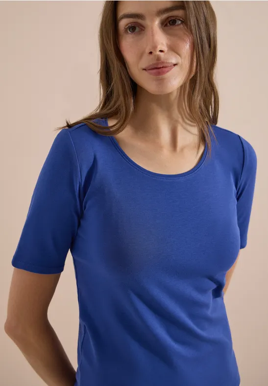 Vrouw in strak blauw T-shirt, zacht glimlachend tegen neutrale achtergrond.