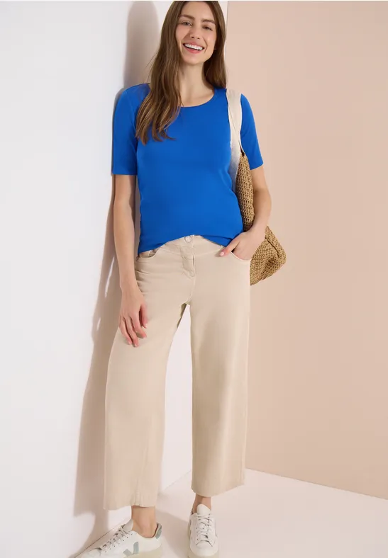 Vrouw leunt tegen muur, draagt blauwe top, beige wijdvallende broek, witte sneakers en rieten tas.
