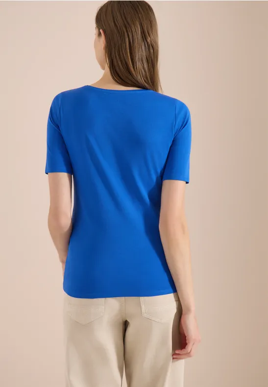 Vrouw gezien van achteren, draagt blauw T-shirt en lichte broek tegen neutrale achtergrond.