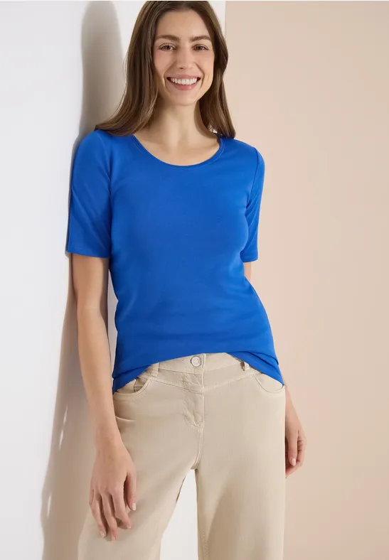 Een vrouw draagt een blauw t-shirt en beige broek, staand voor een lichte achtergrond.