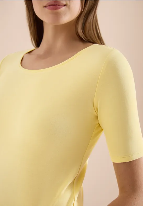 Frau trägt gelbes kurzärmliges Shirt, obere Körperhälfte vor hellem Hintergrund.