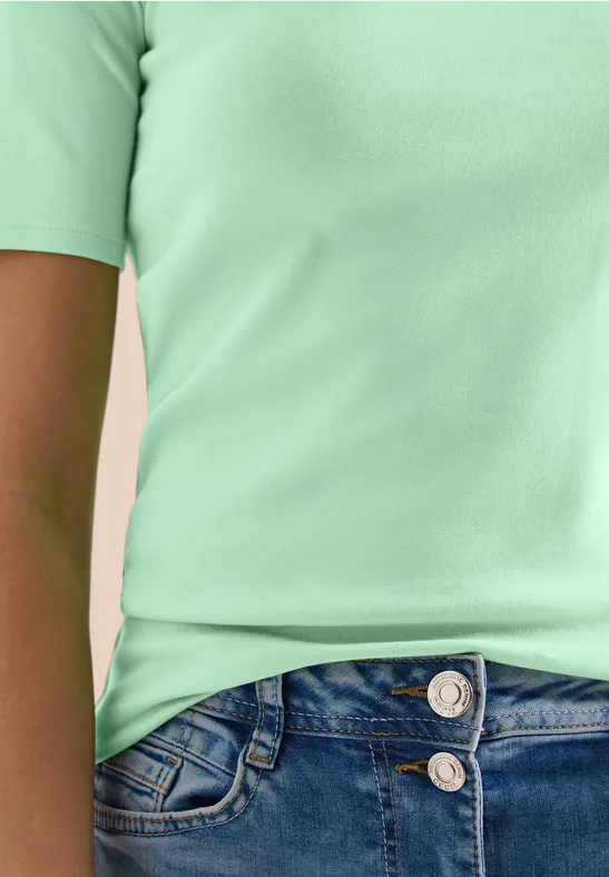 Oberkörper einer Person in mintgrünem T-Shirt und blauen Jeans, zwei Knöpfe sichtbar.