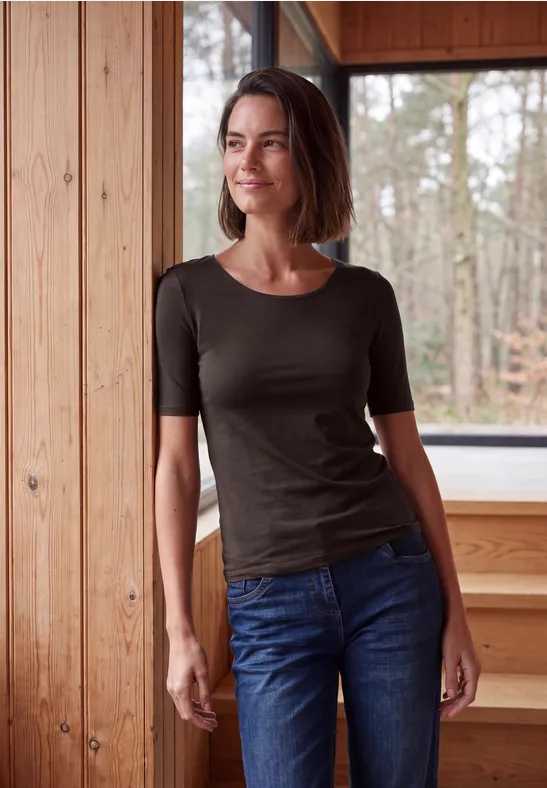Frau lehnt entspannt an hölzernen Wand, Blick auf den Wald, sie trägt Jeans und ein dunkles Shirt.