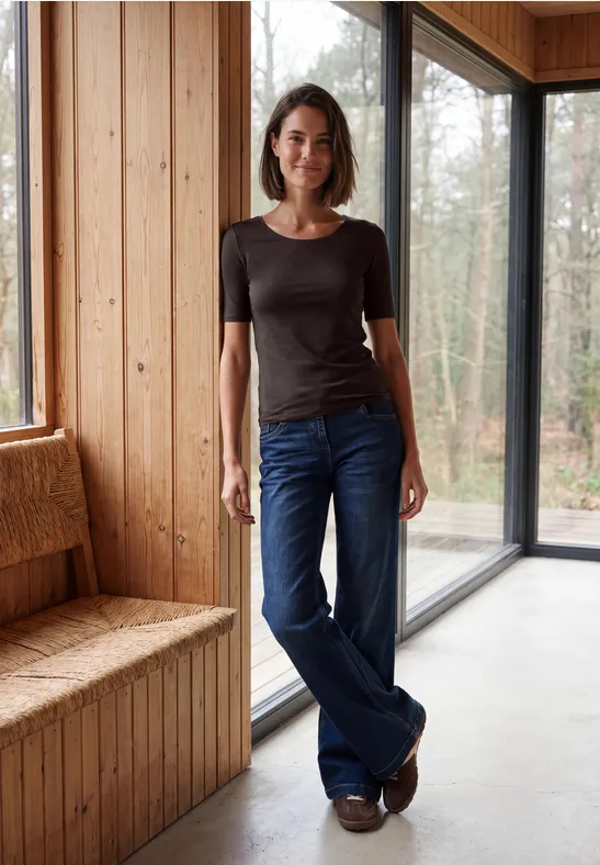 Eine junge Frau in Jeans und T-Shirt lehnt entspannt am Fenster eines modernen Holzinterieurs.