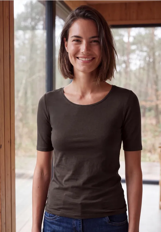 Lächelnde Frau in dunkelgrauem T-Shirt und Jeans steht in Holzveranda vor großen Fenstern.