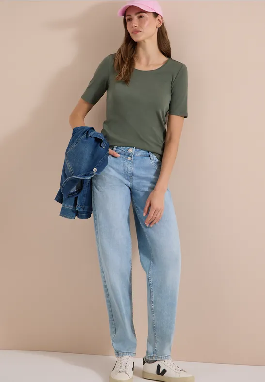Frau in olivgrünem Shirt, weiten hellblauen Jeans, weißen Sneakers, mit Jeansjacke über dem Arm.