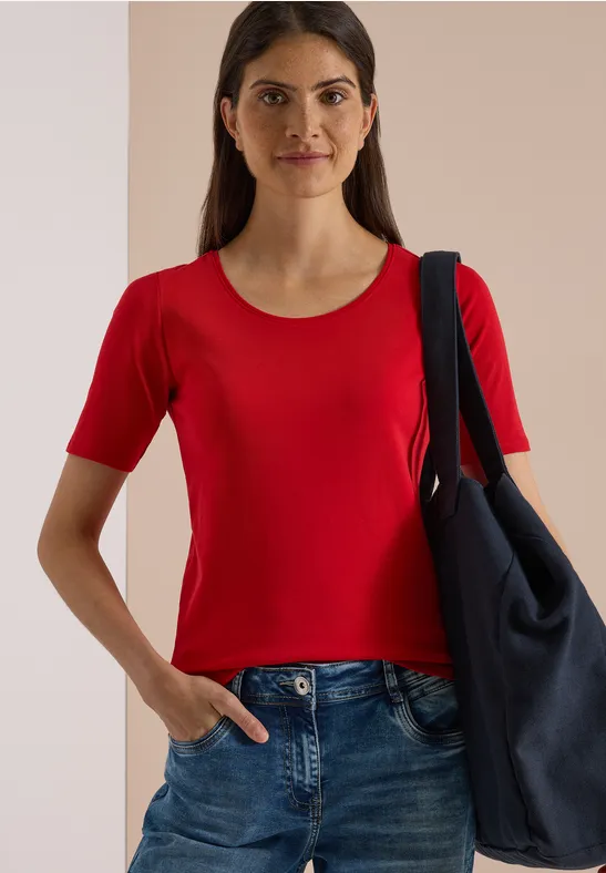 Frau in rotem Kurzarmshirt, Jeans und großer Tasche über der Schulter vor hellem Hintergrund.