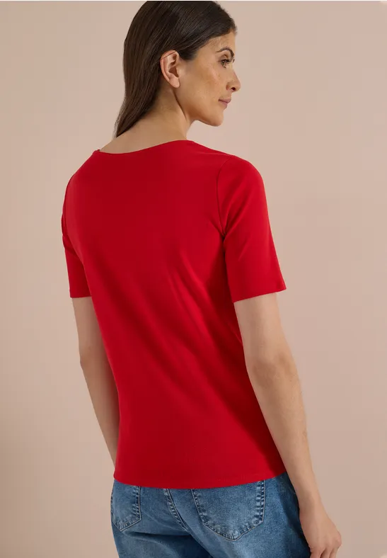 Eine Frau von hinten in rotem T-Shirt und blauer Jeans vor neutralem Hintergrund.