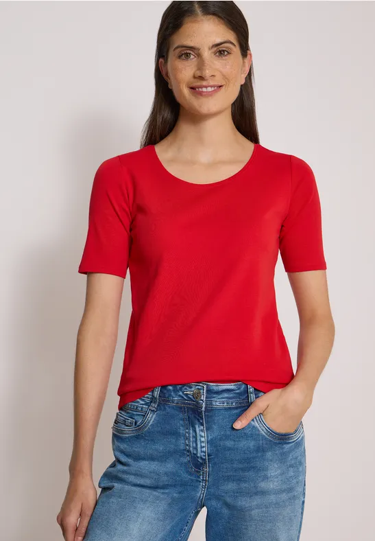 Frau mit rotem T-Shirt und blauer Jeans, lächelnd vor hellem Hintergrund.