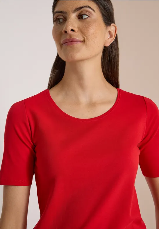 Frau mit rotem T-Shirt mit kurzen Ärmeln und rundem Ausschnitt, Blick leicht seitlich.