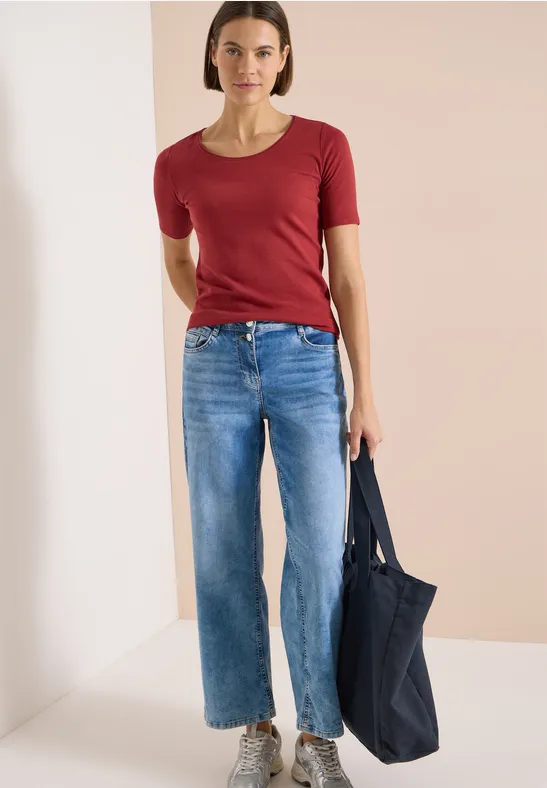 Frau trägt rotes T-Shirt, blaue Jeans und hält eine dunkelblaue Tasche.