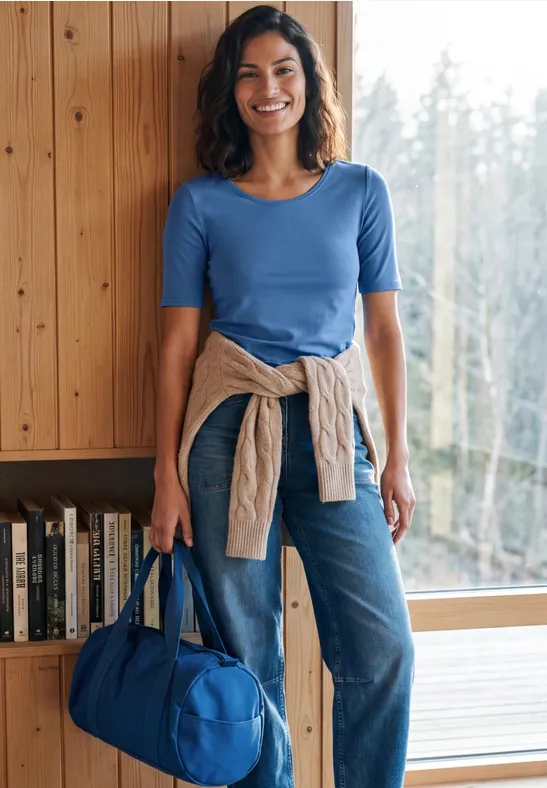 Lächelnde Frau in blauem Shirt und Jeans, hält eine blaue Tasche vor Fenster und Bücherregal.