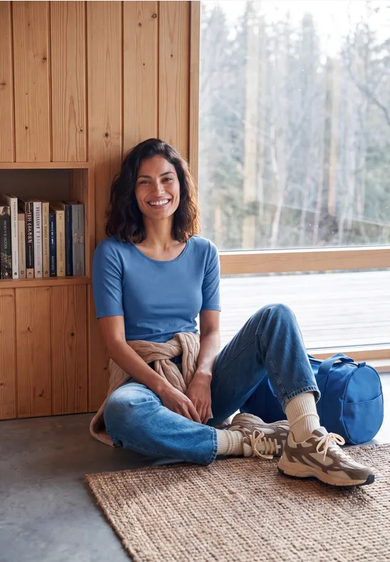 Lächelnde Frau sitzt im Holzhaus am Fenster, trägt Jeans, blaues Shirt und Wanderschuhe.