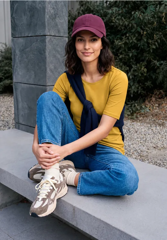 Junge Frau in gelbem T-Shirt, Jeans und Mütze sitzt lächelnd im Freien auf einer Steinbank.