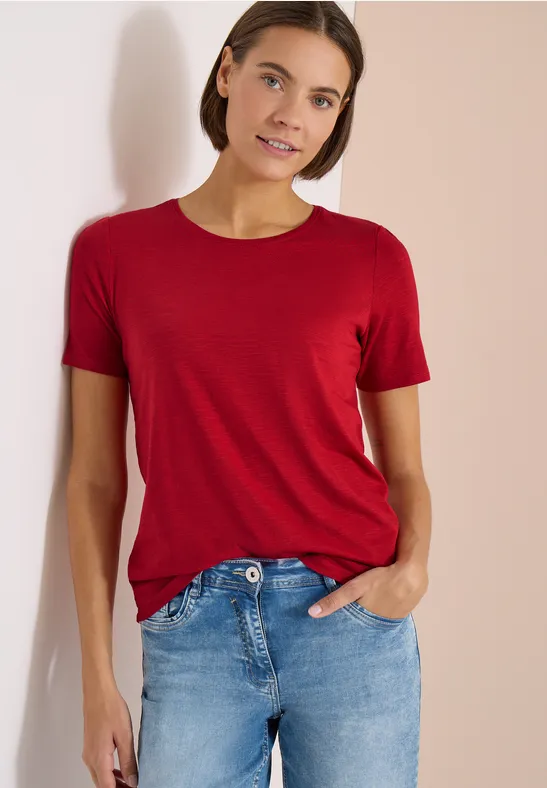 Frau in rotem T-Shirt und blauen Jeans vor hellem Hintergrund.