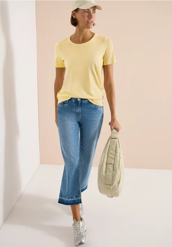 Vrouw in geel t-shirt, jeans en pet met een tas en sneakers.