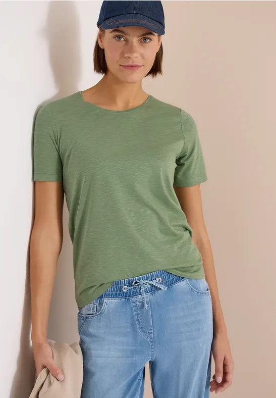 Person in grünem T-Shirt und hellblauen Jeans lehnt an einer Wand.