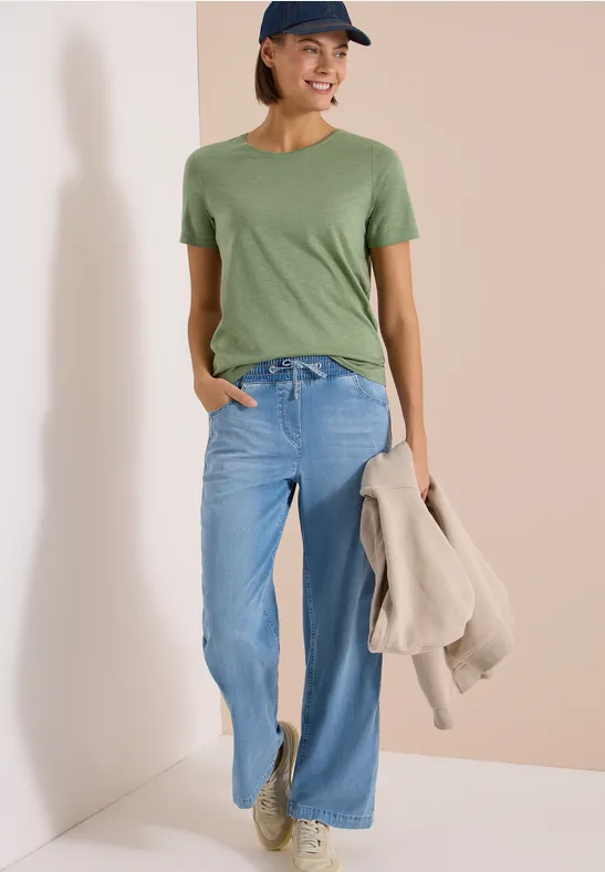 Frau in grünem T-Shirt, weiten Jeans und Basecap, lässig stehend, trägt Jacke in der Hand.