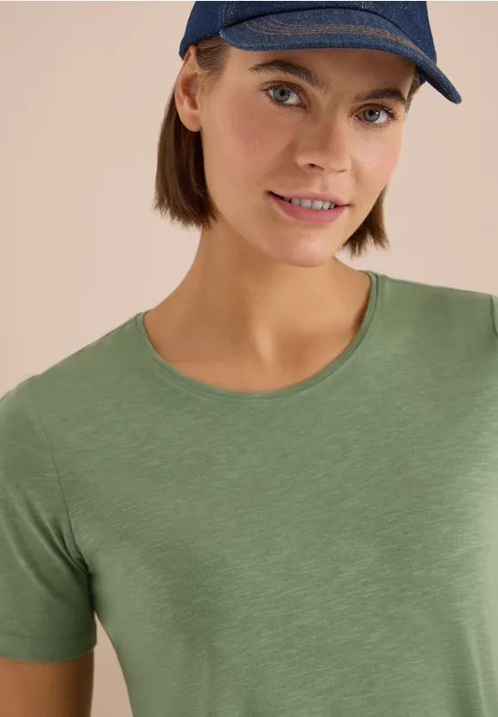 Person mit grünem T-Shirt und blauem Cap vor neutralem Hintergrund.