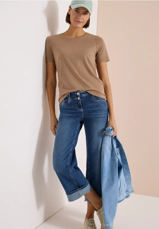 Vrouw geleund tegen muur, bruine t-shirt, blauwe jeans, spijkerjasje in hand.