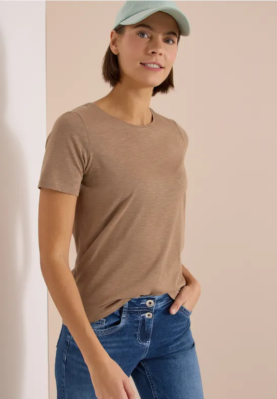 Persoon met bruin T-shirt, blauwe jeans en groene pet, staand in zijaanzicht met één hand in de zak.