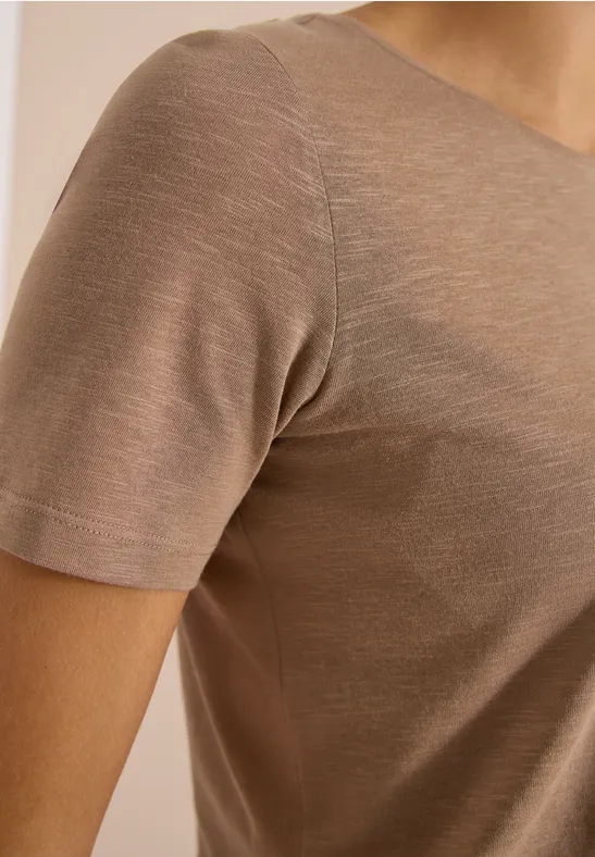 Close-up van een persoon in een beige/bruin, nauwsluitend T-shirt met korte mouwen van dunne stof.