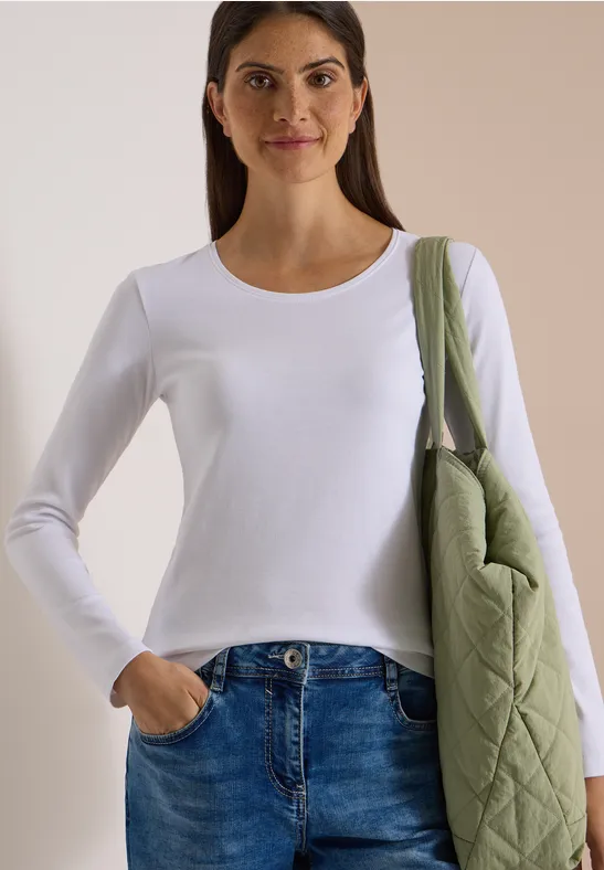 Eine Frau in weißem Langarmshirt und Jeans, mit grünem Stepp-Schulterbeutel vor neutralem Hintergrund.