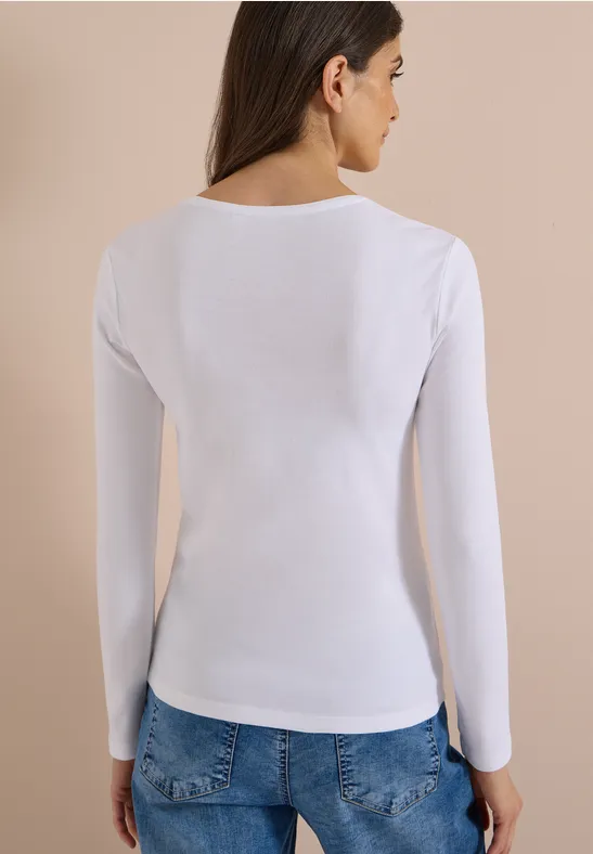 Rückansicht einer Frau in weißem langärmligem Oberteil und blauen Jeans vor neutralem Hintergrund.