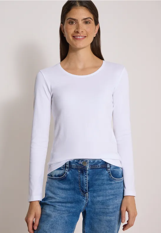 Eine Frau in weißem Langarmshirt und Jeans vor hellem Hintergrund.