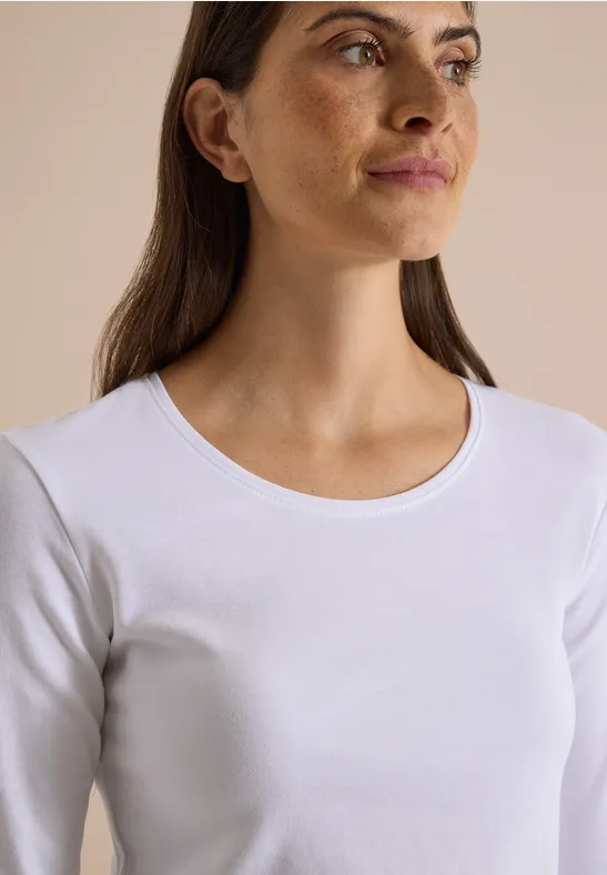 Junge Frau mit braunen Haaren, im weißen Langarmshirt vor neutralem Hintergrund, seitlich blickend.