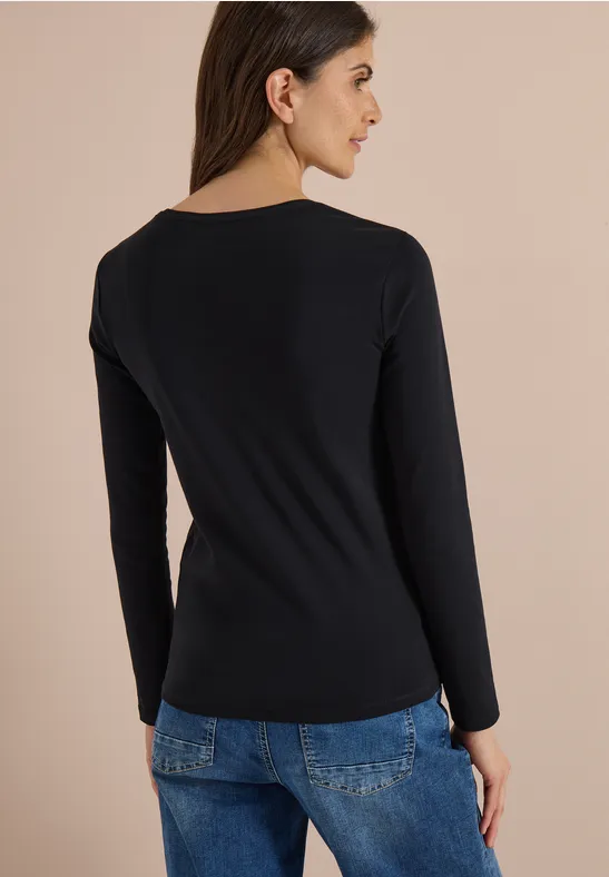 Frau von hinten in schwarzem Langarmshirt und blauen Jeans vor beigem Hintergrund.