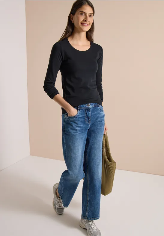 Frau in schwarzem Langarmshirt, blauen Jeans und Sneakern hält eine grüne Tasche und lächelt.
