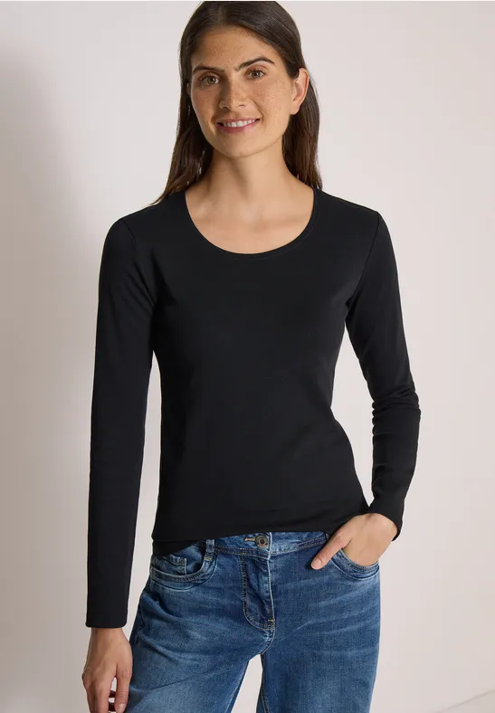 Frau mit schwarzem Langarmshirt und blauen Jeans lächelt vor hellem Hintergrund.