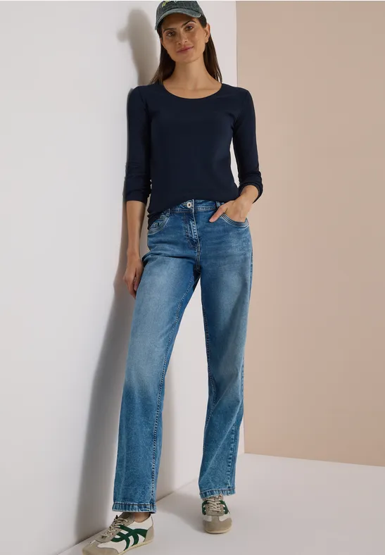 Eine Frau lehnt entspannt an einer Wand und trägt ein dunkelblaues Shirt, Jeans, Baseballcap und Turnschuhe.