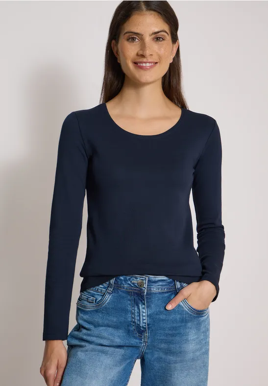 Eine lächelnde Frau trägt eine dunkelblaue Langarmbluse und eine blaue Jeans vor neutralem Hintergrund.