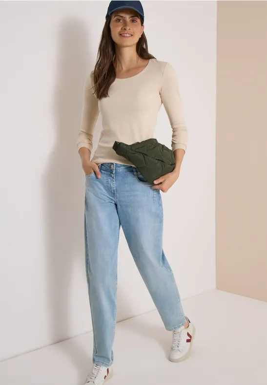 Frau in Jeans, weißem Langarmshirt, Cap und Umhängetasche lehnt an heller Wand.