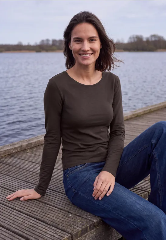 Een glimlachende vrouw zit ontspannen op een houten steiger aan een meer in casual kleding.