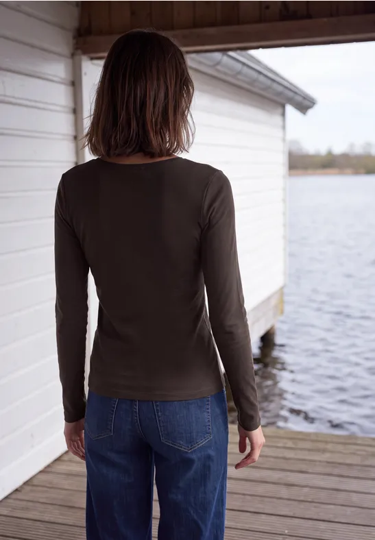 Een persoon in donker shirt met lange mouwen en jeans staat op een steiger bij een boothuis en kijkt naar het water.