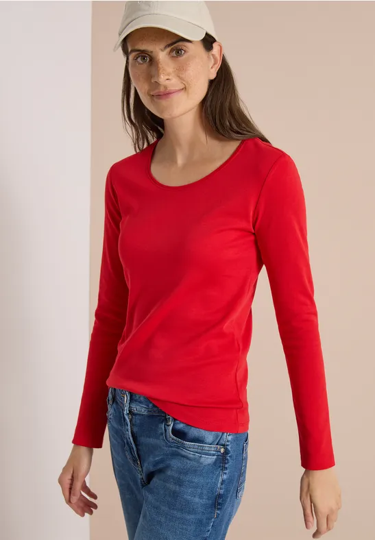 Eine Frau mit rotem Langarmshirt, blauen Jeans und beige Kappe vor neutralem Hintergrund lächelt sanft.