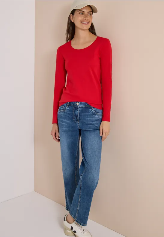 Frau mit rotem Langarmshirt, blauen Jeans, weißen Sneakern und Basecap steht lächelnd in heller Ecke.