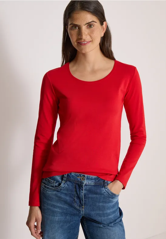 Frau trägt rotes Langarmshirt und Jeans, steht mit Hand in Tasche und lächelt.