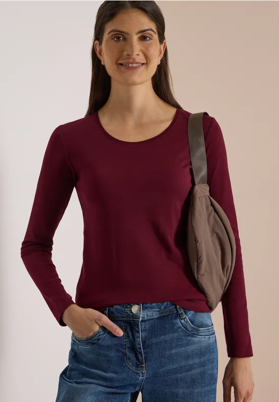 Frau in dunkelrotem Langarmshirt, blauen Jeans und Schultertasche, die lächelnd vor beiger Wand steht